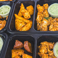 Black meal prep containers filled with seasoned roasted potato wedges, grilled chicken pieces, and small containers of green sauce, arranged in a grid pattern.