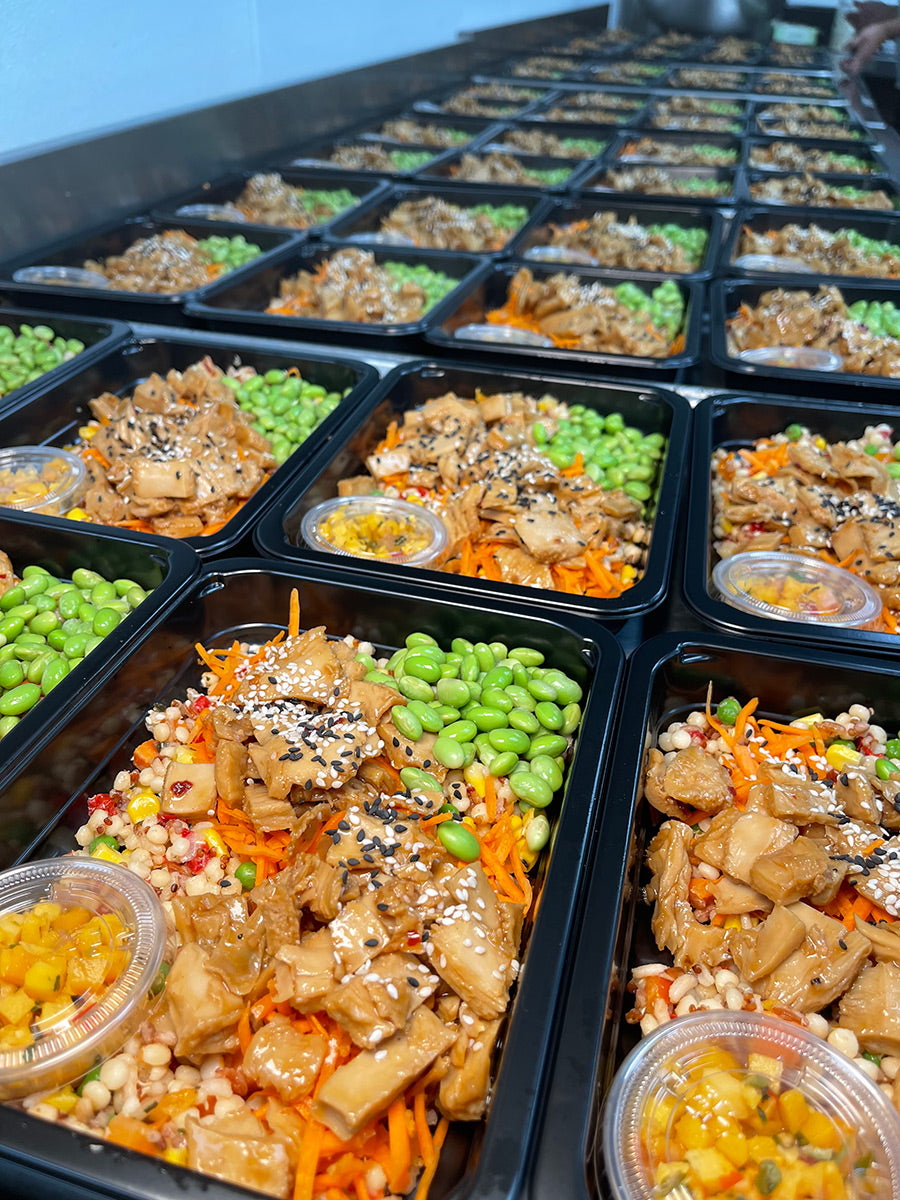 Rows of black meal prep containers filled with sesame chicken, shredded carrots, edamame, pearl couscous, and small cups of yellow sauce, arranged neatly on a countertop.