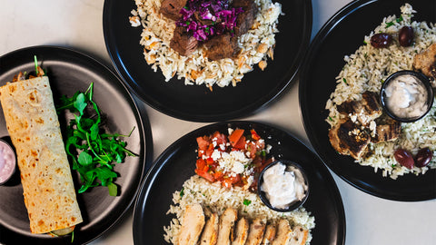 Four black plates with Mediterranean dishes: one with a wrap and greens, two with rice, grilled meat, and tzatziki, and one with rice, chicken, tomato salad, feta, and tzatziki. All plates are arranged on a white table.