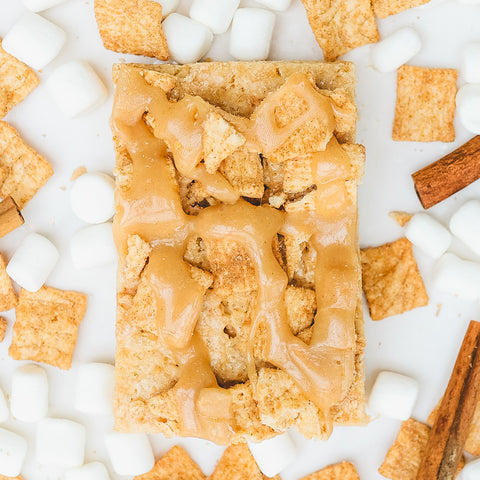 A Cinna Krunch Buff Krispy Treat (Subscription) topped with cinnamon cereal pieces and caramel drizzle, surrounded by mini marshmallows, extra cereal squares, and cinnamon sticks on a white surface.