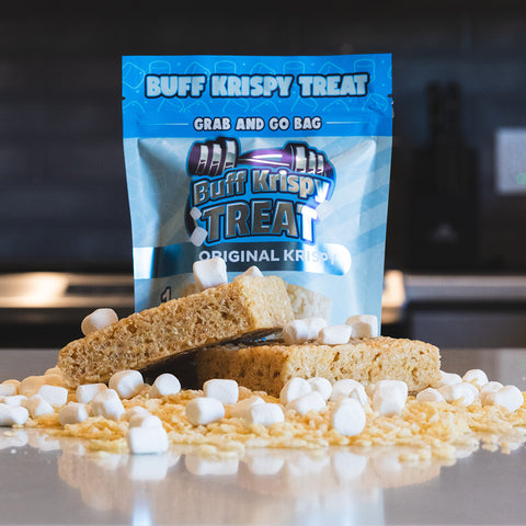 A bag of Buff Krispy Treat stands behind three Buff Krispy Treat bars, with mini marshmallows and crispy rice cereal scattered on a kitchen counter.
