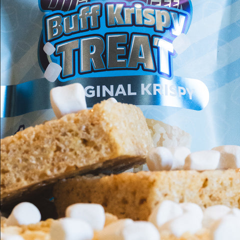 Close-up of Buff Krispy Treat bars stacked in front of the package, with marshmallows scattered around. Buff Krispy Treat is a delicious, low-calorie protein bar ideal for muscle recovery.