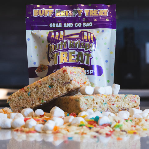 A Fruity Cereal Buff Krispy Treat stands behind two large rice crispy treats, surrounded by mini marshmallows and colorful cereal pieces—a delicious treat made with cereal milk whey protein isolate.