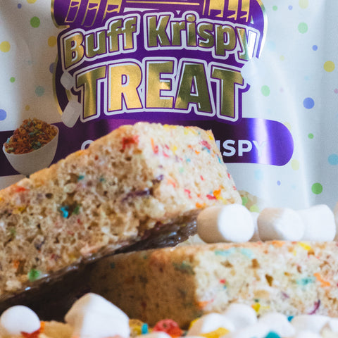Close-up of two Fruity Cereal Buff Krispy Treats with mini marshmallows in front, and a festive Buff Krispy Treat package in the background—a high protein snack made with cereal milk whey protein isolate.