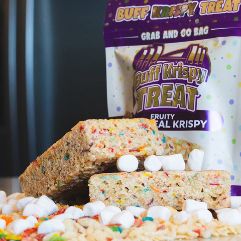 Two large Fruity Cereal Buff Krispy Treats, made with cereal milk whey protein isolate, are stacked in front of their branded bag. Mini marshmallows and cereal pieces are scattered on the table around them.