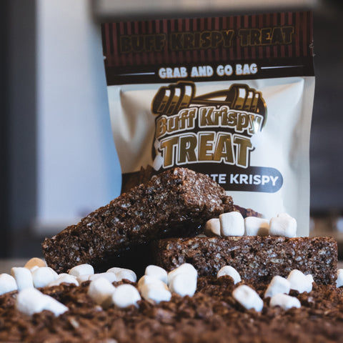 A close-up of chocolate crispy treats stacked with mini marshmallows, with a package labeled Chocolate Buff Krispy Treat in the background, highlighting this guilt-free snack.