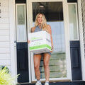 A smiling woman in athletic wear stands on a porch, holding a green and white box labeled “Herbalife Fit” in front of a glass door on a white house.
