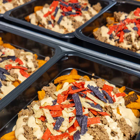 Black trays filled with food featuring seasoned ground beef, creamy sauce, crispy tortilla strips in red, purple, and white, served over a bed of sweet potato fries or wedges.