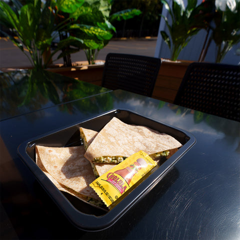 A black tray with quesadilla slices and a packet of Cholula hot sauce sits on a shiny outdoor table, with green plants and empty chairs in the background.