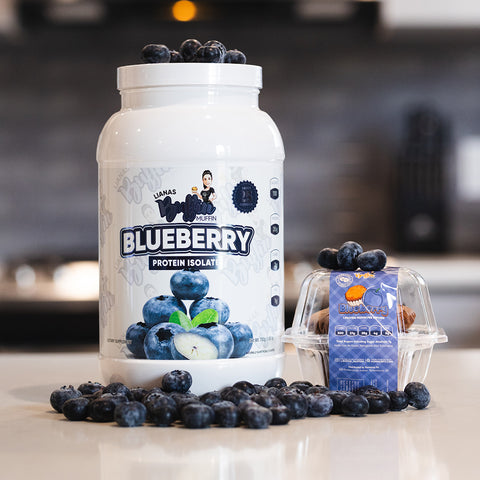 A large container of Liana's Buffin Muffin - Protein Isolate and a small plastic box of fresh blueberries are on the counter with some loose berries around them, set against a blurred kitchen background.
