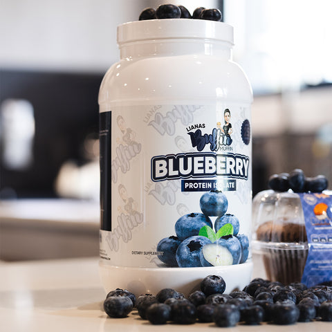 A large white container labeled "Liana's Buffin Muffin - Protein Isolate" with images of blueberries sits on a counter, surrounded by fresh blueberries and a packaged muffin—a great choice for dessert lovers and whey protein fans.