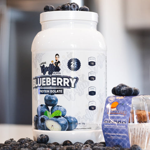 A white tub of Liana's Buffin Muffin - Protein Isolate with blueberry dessert-inspired flavors sits on a kitchen counter, featuring blueberries on the label and surrounded by fresh blueberries and a small plastic fruit container.