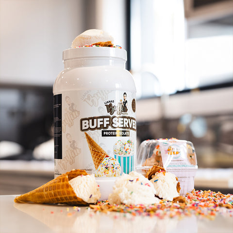 A large white container of Lianas Buffin Muffin - Protein Isolate with ice cream cone graphics sits on a counter, surrounded by dessert-inspired flavors, waffle cones, and colorful sprinkles in a bright kitchen.