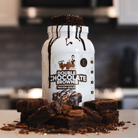 A tub of Lianas Buffin Muffin - Protein Isolate (Subscription) sits on a counter, topped with stacked brownies drizzled in chocolate, surrounded by more brownies and crumbs, with a blurred kitchen visible in the background.