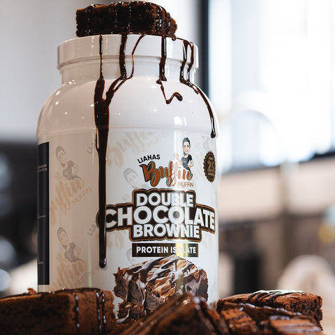 A container of Liana's Buffin Muffin - Protein Isolate is topped with brownies and drizzled with chocolate sauce, with more brownies stacked nearby in a bright, modern kitchen.