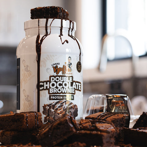 A large container of Lianas Buffin Muffin - Protein Isolate rests on the counter, surrounded by rich brownies with chocolate syrup, highlighting its indulgent dessert-inspired flavor in a bright kitchen scene.