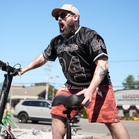 A bearded man in sunglasses, a beige cap, and the Men’s Mesh Jersey – Black White Trim stands by an electric scooter, shouting or singing with his mouth wide open on a sunny day.