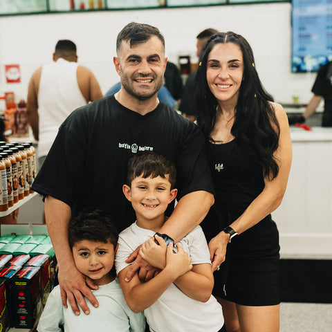 A smiling family of four poses together in a store. The parents stand behind their two young children, hugging them. Shelves with products are visible, and other people are in the background.