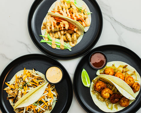 Three black plates, each with a taco and a sauce container. The tacos have different fillings: grilled chicken with sauce, shrimp with sauce, and shredded cheese with meat. A lime wedge is next to the shrimp taco, all on a white marble surface.