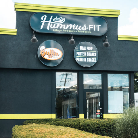 A gray building with green trim featuring signs for Hummus Fit and Buffin advertising meal prep, protein shakes, and protein snacks, with bushes in front and windows reflecting the street.