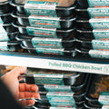 A hand reaching for a packaged meal labeled Pulled BBQ Chicken Bowl from a shelf stacked with various ready-to-eat chicken bowls in plastic containers.