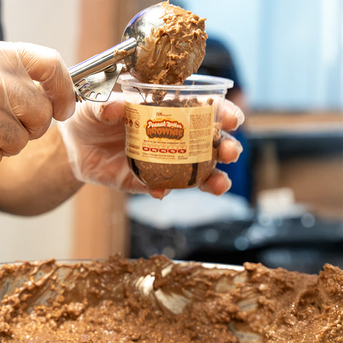 A gloved hand uses an ice cream scoop to fill a plastic cup labeled Peanut Butter Brownie with thick, brown dessert batter. A large container of the batter is visible in the foreground.