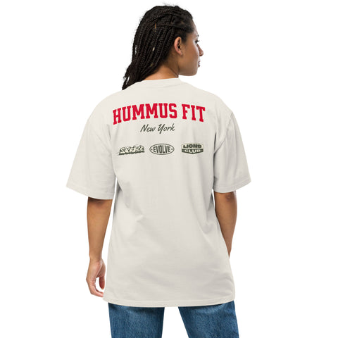 A person with braided hair wears a white T-shirt with HUMMUS FIT New York in red and black text, and several logos printed on the back. The person faces away from the camera.