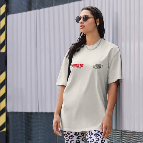 A woman wearing sunglasses and a light-colored T-shirt with the words HUMMUS FIT and EVOLVE stands against a metallic fence. She has braided hair and is also wearing patterned pants and a chain necklace.