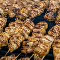 Close-up of several skewers of grilled, seasoned chicken cooking on a barbecue grill. The chicken pieces are golden brown with visible grill marks.
