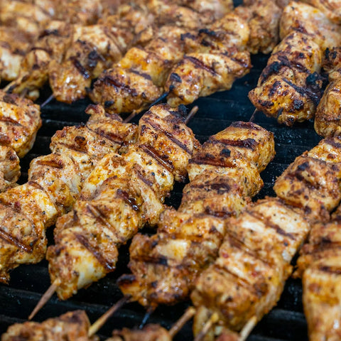 Close-up of several skewers of grilled, seasoned chicken cooking on a barbecue grill. The chicken pieces are golden brown with visible grill marks.