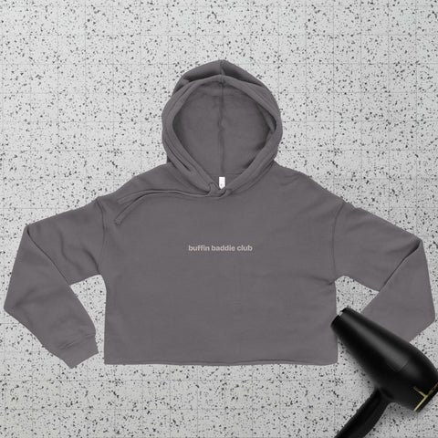 A gray cropped hoodie with the words buffin baddie club printed on the front lies on a speckled surface. A black hair dryer is partially visible in the lower right corner.