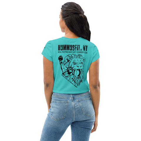 A woman with long dark hair wears the Turquoise Hummus Fit Script Crop Tee with an all-over Statue of Liberty eating design and HUMMUSFIT, NY Meal Prepping Made Easy – hummusfit.com text on the back. She pairs it with blue jeans.