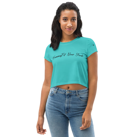 A woman stands facing forward against a plain white background, smiling softly at the camera in a Turquoise Hummus Fit Script Crop Tee paired with blue jeans.