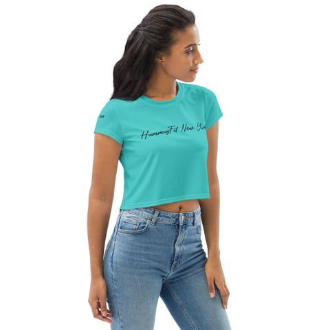 A woman stands against a plain white background, looking to her left, wearing a Turquoise Hummus Fit Script Crop Tee with four-way stretch fabric, paired with light blue jeans.