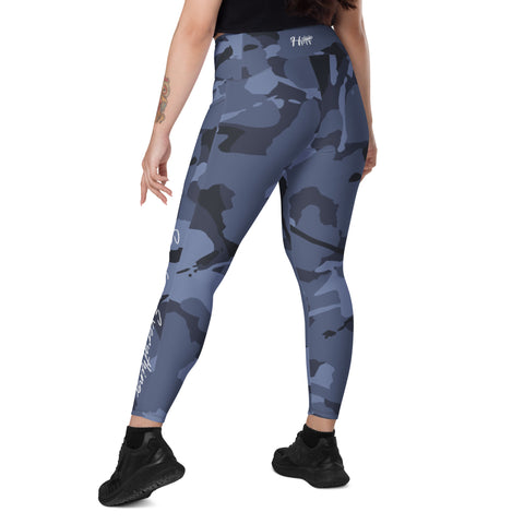 A person with a left arm tattoo, wearing Blue Camo Leggings With Pockets and black sneakers, faces away to show the leggings’ back. The outfit features a high waist and dark blue/black camo print, set against a plain white background.