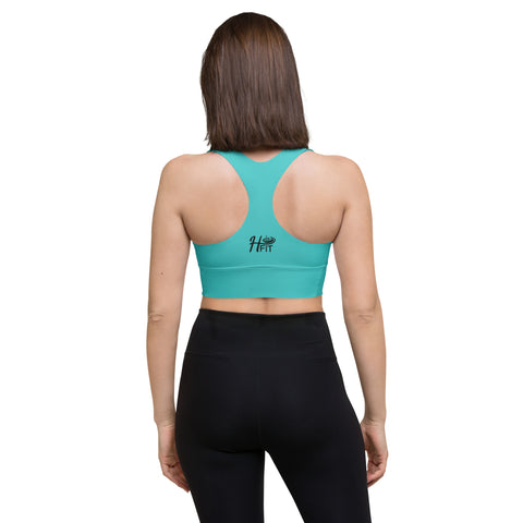 A woman with straight brown hair faces away, wearing the Hummus New York Sports Bra in turquoise, featuring the HF Fit logo on the back and high-waisted black leggings, standing against a white background.
