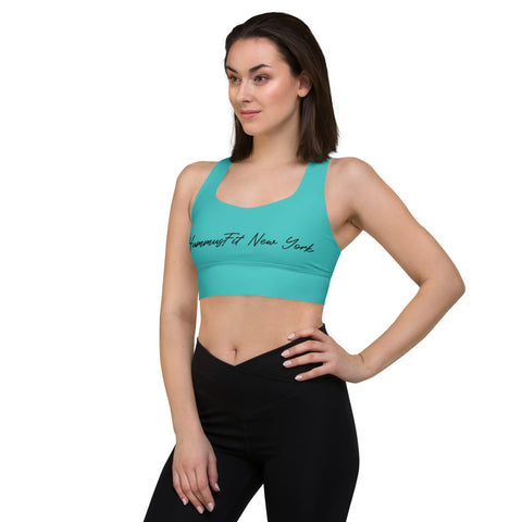 A woman stands with one hand on her hip, looking to the side, in a turquoise Hummus New York Sports Bra and black leggings. The sports bra features soft compression fabric and is set against a plain white background.