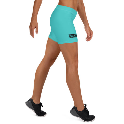 A woman in a side-walking pose wears the Womens Turquoise Shorts featuring a comfy elastic waistband and HUMM print on the left leg, paired with a matching sports bra and black athletic shoes on a white background.