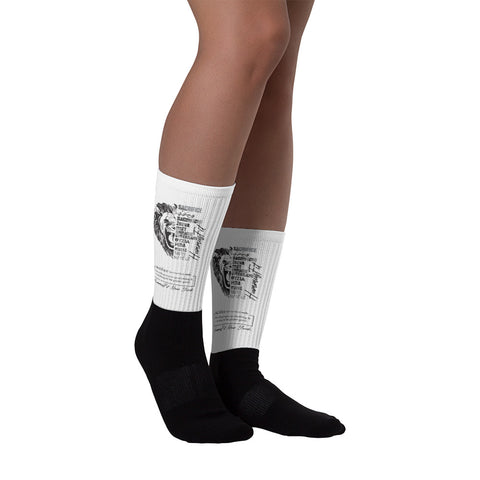 Someone stands on a white surface wearing Sacrifice Worldwide Socks—comfortable, knee-high socks in white with black soles and a black printed design on the sides.