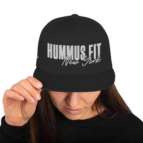 A person wearing a Black Snapback Hat with white HUMMUS FIT New York text holds the brim, partially covering their face.