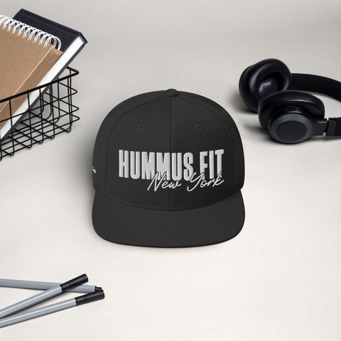 The Black Snapback Hat, featuring HUMMUS FIT New York embroidered in white, rests on a beige surface beside a wire basket holding notebooks, black headphones, and two pens.