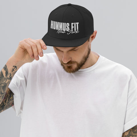 A man with tattoos, wearing a plain white t-shirt and a Black Snapback Hat that reads HUMMUS FIT New York, looks downward while holding the hat’s brim against a plain gray background.