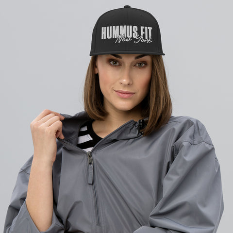 A woman poses against a plain, light gray background wearing a gray jacket and a Black Snapback Hat.