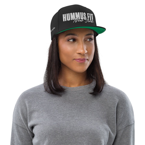 A woman in a gray sweater wears the Black Snapback Hat, featuring an adjustable fit and HUMMUS FIT New York in white and silver lettering, with a green underbill, looking to the side with a neutral expression.
