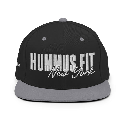 The Black & Silver Snapback Hat features HUMMUS FIT New York embroidered in bold white and script on the front, offering a classic adjustable fit for any style.