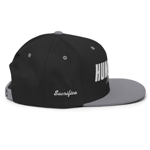 The Black & Silver Snapback Hat features “Sacrifice” embroidered in white on the side and part of “HUM” boldly displayed on the front, offering a classic fit for everyday wear.