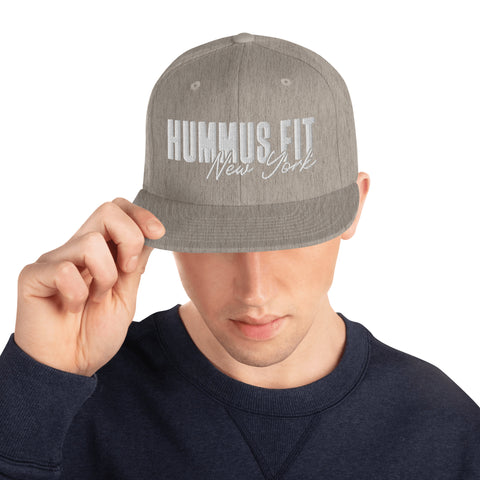 A person wears the Heather Snapback Hat, a gray classic fit cap with white HUMMUS FIT New York text, tilting its flat brim down. The adjustable snap closure hat is paired with a dark sweatshirt.