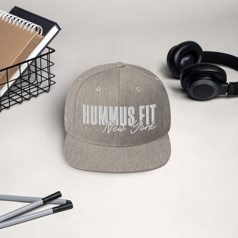 The Heather Snapback Hat, a gray flat-bill cap with HUMMUS FIT New York on the front and an adjustable snap closure, is displayed on a light surface beside black headphones, pens, and a wire basket holding notebooks.