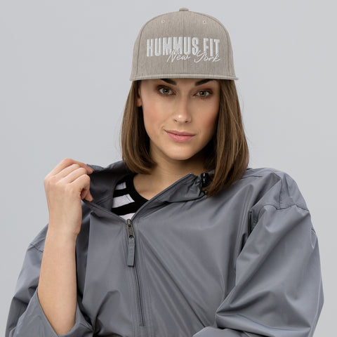A woman in a gray jacket and striped shirt wears the Heather Snapback Hat with a flat brim, looking at the camera with a neutral expression while holding her jacket’s collar against a plain light gray background.