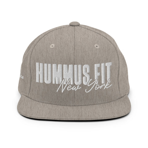 The Heather Snapback Hat is a light gray classic fit with a flat brim and “HUMMUS FIT New York” embroidered in large white letters. It features an adjustable snap closure and is shown facing forward against a white background.